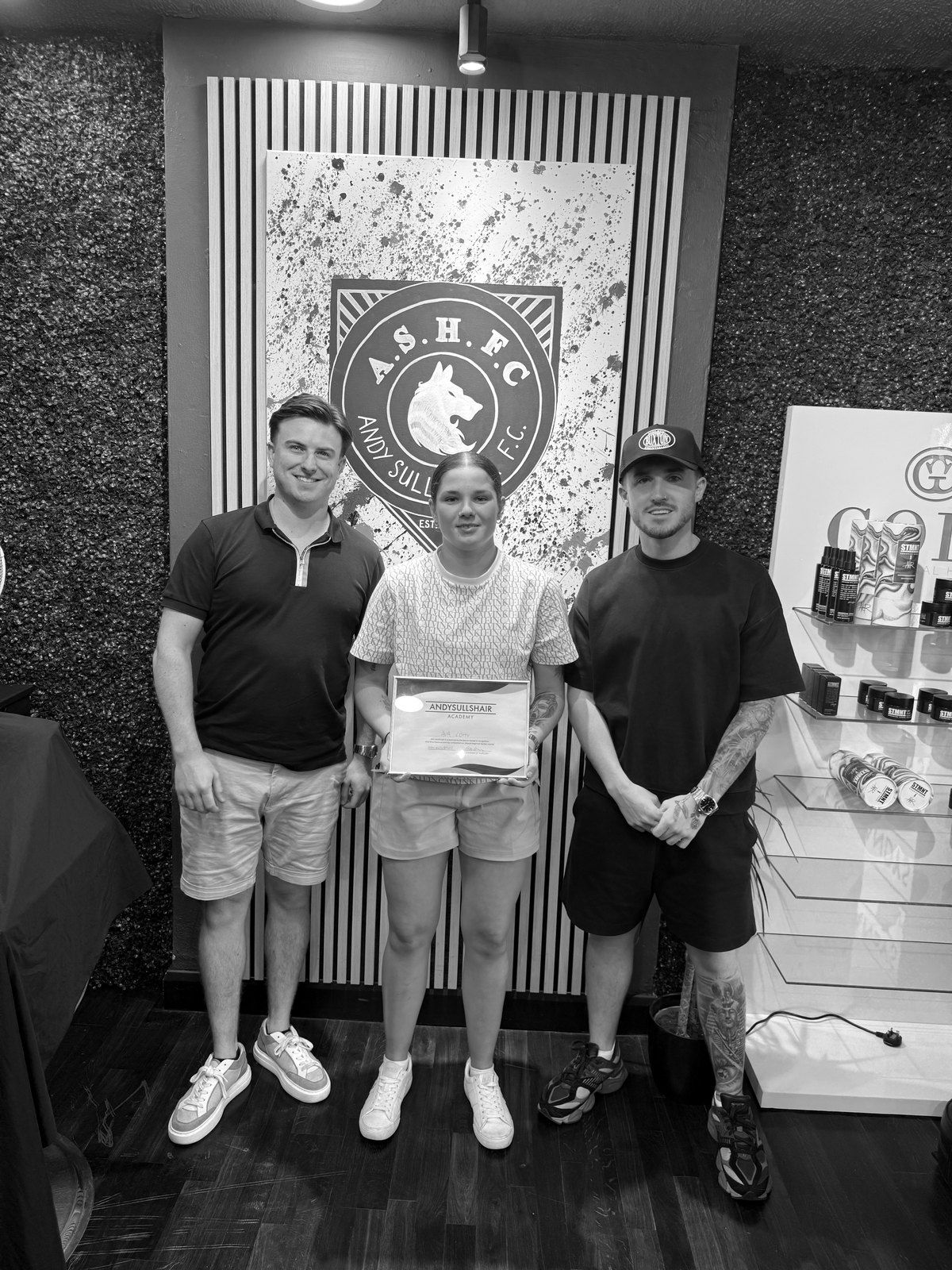 Andy with two graduates holding their AndySullsHair Academy certificates in front of the crest wall