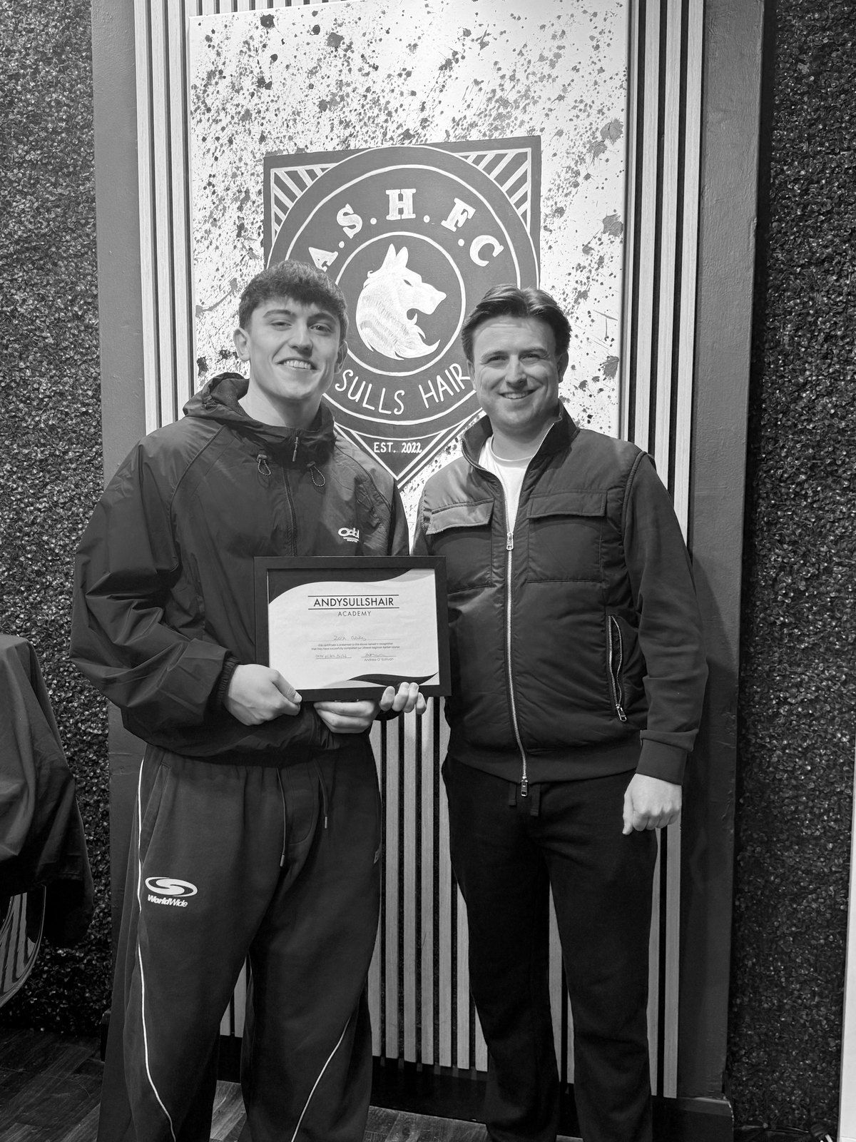 Andy with a graduate and his certificate in front of the crest wall